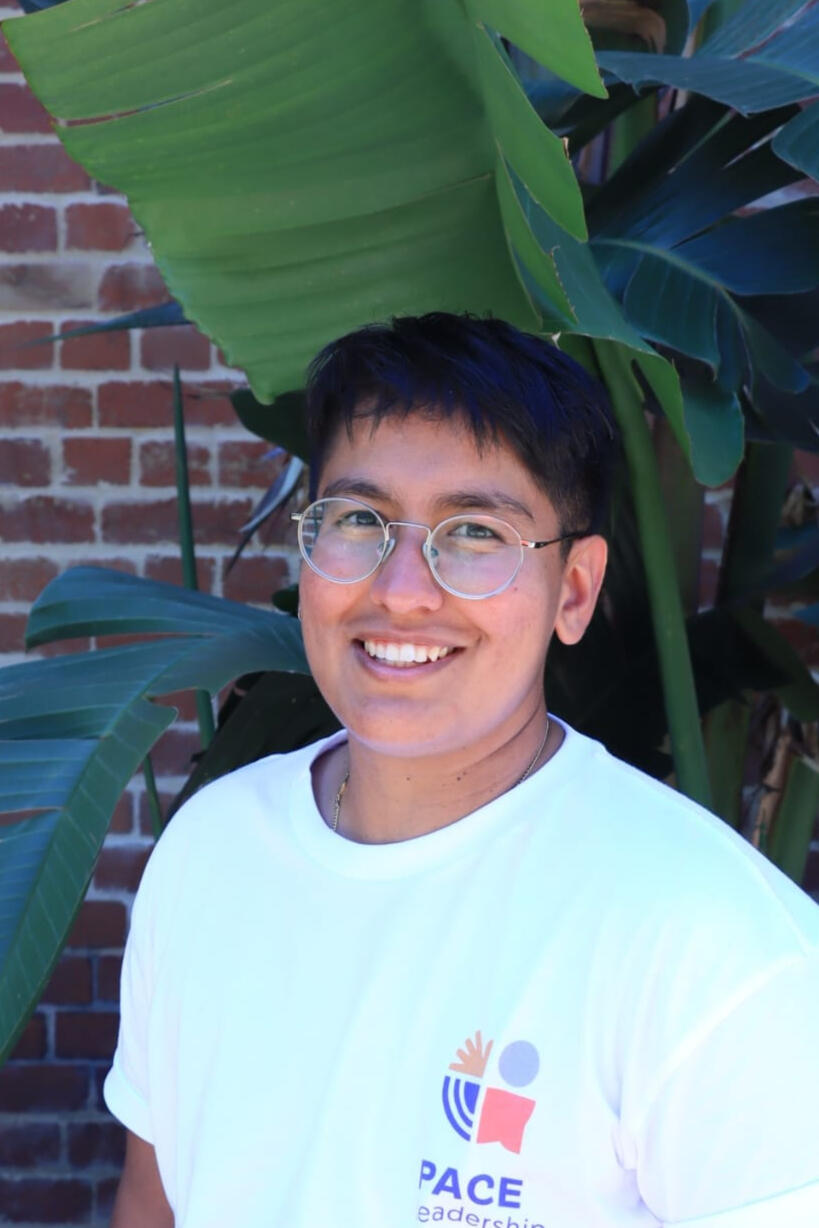A person with short hair, medium skin tone and wire rimmed glasses wearing a white shirt and standing by a brick wall